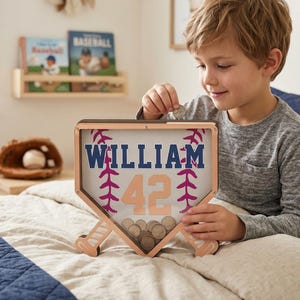 Puede incluir: Una caja de sombra de b&eacute;isbol de madera con el nombre "WILLIAM" y el n&uacute;mero "42" en azul marino y melocot&oacute;n. La caja tiene forma de plato de home y tiene detalles de bate de b&eacute;isbol. La caja est&aacute; llena de monedas.