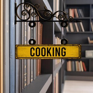 May include: A yellow rectangular sign with the word "COOKING" in bold black letters hangs from a decorative black metal bracket. The sign is suspended by chains and set against a blurred background of bookshelves.