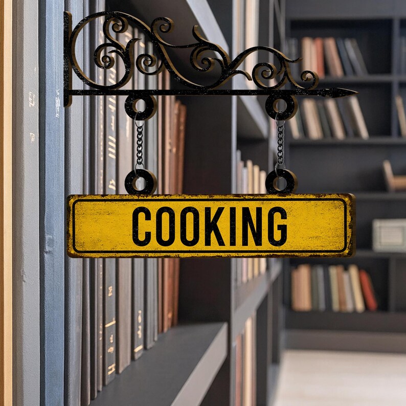 May include: A yellow rectangular sign with the word "COOKING" in bold black letters hangs from a decorative black metal bracket. The sign is suspended by chains and set against a blurred background of bookshelves.