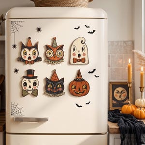 Può includere: Un set di sei magneti a tema Halloween raffiguranti un gatto, un gufo, un fantasma, un teschio, un gatto nero e una zucca, tutti con cappellini da festa o papillon, applicati su un frigorifero color crema decorato con adesivi di pipistrelli.