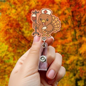 May include: A badge reel featuring a cartoon turkey wearing a nurse's cap and holding a pumpkin spice latte. The turkey is brown and tan with a red cross on its cap. The IV bag reads "Pumpkin Spice".