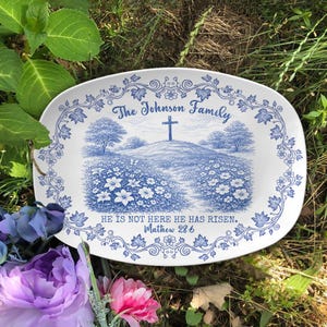 May include: An oval, white ceramic platter with a blue floral border and a central pastoral scene. The scene depicts a cross on a hill, with the text "The Johnson Family" and "HE IS NOT HERE HE HAS RISEN. Matthew 28:6".
