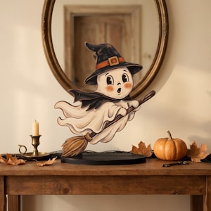 May include: A flat wooden cutout of a cartoon ghost wearing a black witch hat with an orange band, flying on a brown broomstick. The ghost has a white body, rosy cheeks, and a surprised expression. It is mounted on a circular black wooden base.