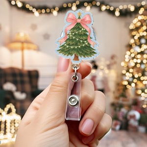 May include: A Christmas tree-shaped badge reel with a pink bow and gold star. The green tree has a light blue outline and is decorated with a string of lights. The reel has a clear plastic clip and a metal retractable cord.