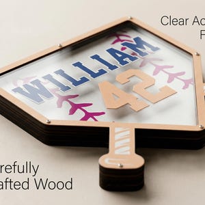 Puede incluir: Una pantalla con tem&aacute;tica de b&eacute;isbol con una parte delantera de acr&iacute;lico transparente y madera cuidadosamente elaborada. El dise&ntilde;o presenta el nombre "WILLIAM" en azul, el n&uacute;mero "42" en beige y costuras de b&eacute;isbol en morado.