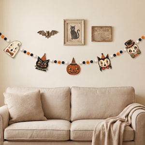 May include: A Halloween garland featuring felt balls in black, white, and orange, interspersed with flat wooden cutouts of a ghost, a black cat, a jack-o'-lantern, a second cat, and a skull, all suspended horizontally against a plain wall.