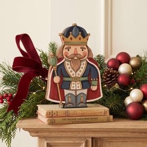 May include: A flat, wooden nutcracker figure wearing a blue jacket, red cape, and gold crown, holding a staff. The figure stands on two stacked vintage books titled The Christmas Book and The Vintage Christmas Books, featuring a painted mustache.