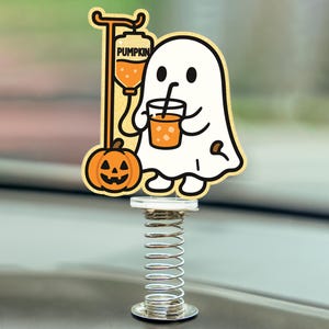May include: A Halloween-themed car accessory featuring a cartoon ghost holding a drink and an IV bag labeled "PUMPKIN". An orange jack-o'-lantern is also present. The image is mounted on a silver spring base.