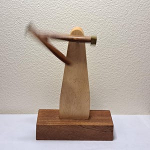 Peut inclure: Une sculpture en bois avec un corps triangulaire de couleur claire sur une base rectangulaire. Un bras en bois brun foncé s'étend du corps, avec une pièce cylindrique de couleur laiton à l'extrémité. La sculpture est posée sur une surface blanche.