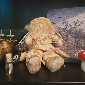 May include: A vintage-style doll with a cream-colored dress, lace collar, and button accents. The doll has curly blonde hair and is seated. A small glass bottle with a cork stopper and a black and white tag are in the foreground. A framed picture of a tree is in the background.