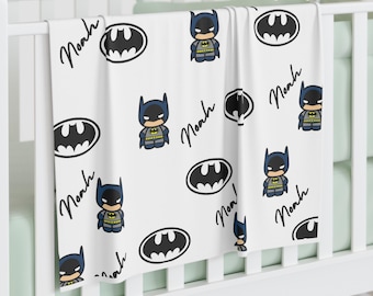 May include: White baby blanket featuring a pattern of Batman logos, cartoon Batman figures, and the name "Noah" in a handwritten style. The blanket is draped over a white crib.