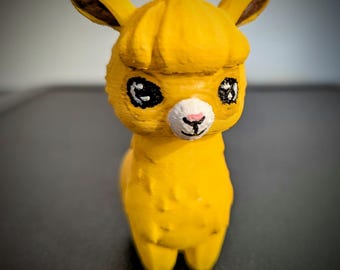 Könnte beinhalten: Eine gelbe Alpaka-Figur mit schwarzen Augen, einer weißen Nase und rosa Nasenlöchern. Das Alpaka hat eine flauschige gelbe Mähne.