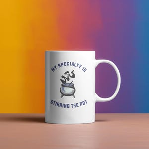 May include: White ceramic mug with the text "MY SPECIALTY IS STIRRING THE POT" in blue. The mug features a graphic of a cauldron with steam rising from it. The mug has a white handle and is set against a colorful background.