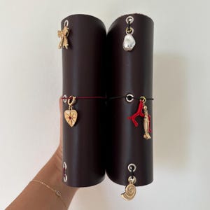 May include: Two dark brown leather journal covers with decorative gold charms. Charms include a bow, a pearl, a heart, a coral branch, a fish, and a seashell. The journals are held together with elastic bands.