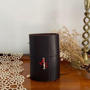 May include: A dark brown leather cylindrical container with a red coral and gold fish charm. The container is on a wooden surface with a lace doily and a brass candlestick in the background.