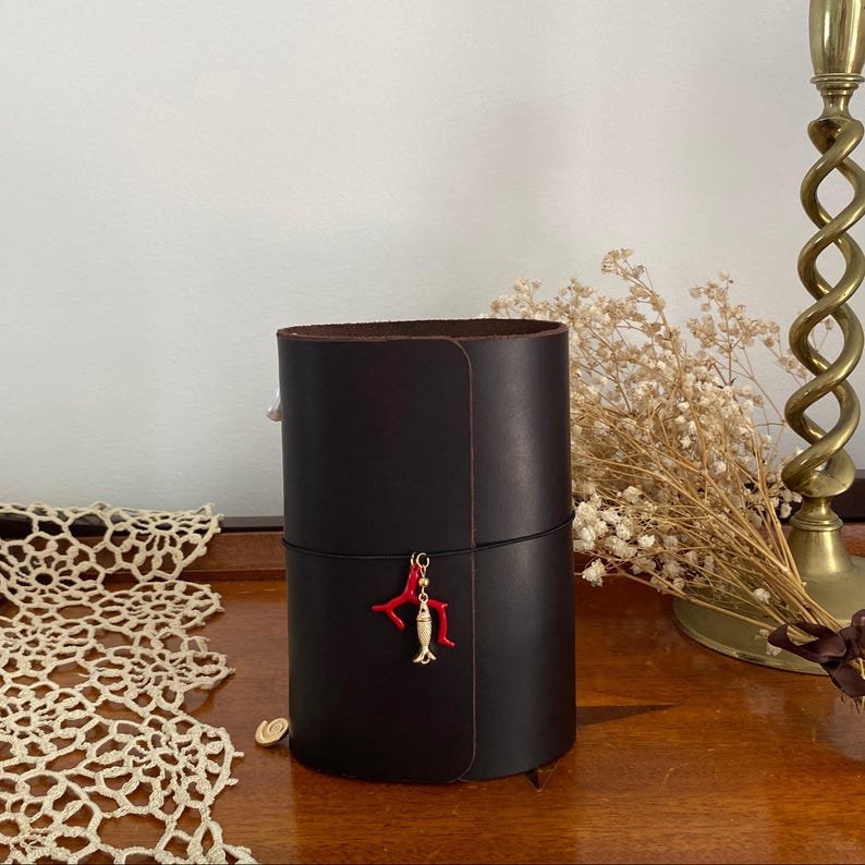 May include: A dark brown leather cylindrical container with a red coral and gold fish charm. The container is on a wooden surface with a lace doily and a brass candlestick in the background.