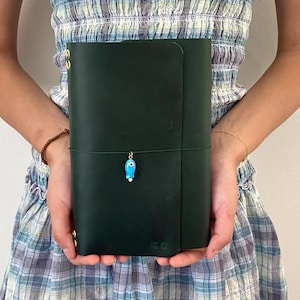 May include: Dark green leather journal with a flap closure and a decorative blue charm. The journal has a string closure and gold-colored metal accents. The initials "KC" are embossed on the lower right corner.