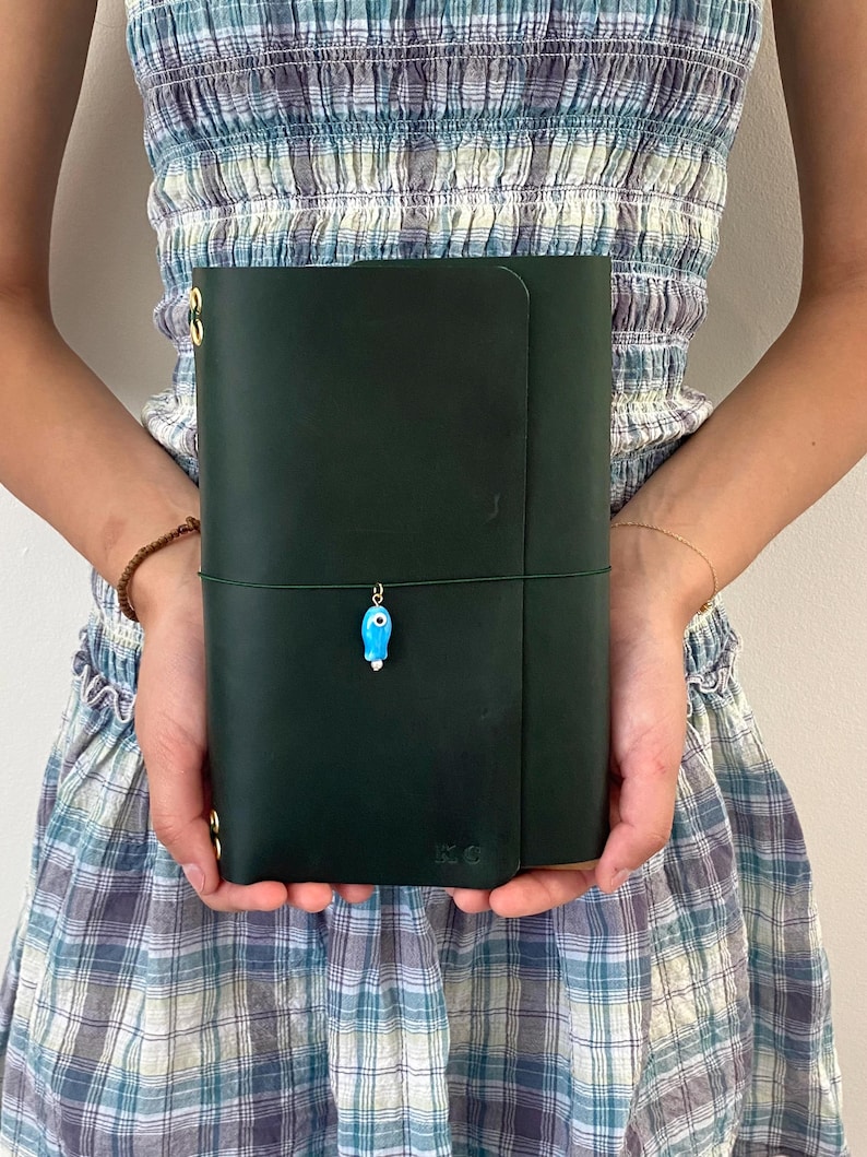 May include: Dark green leather journal with a flap closure and a decorative blue charm. The journal has a string closure and gold-colored metal accents. The initials "KC" are embossed on the lower right corner.