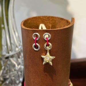 May include: A brown leather cuff with two silver eyelets and a gold star charm. The star charm is embellished with small, clear stones. The cuff is stitched with pink thread.