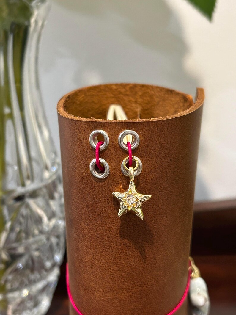 May include: A brown leather cuff with two silver eyelets and a gold star charm. The star charm is embellished with small, clear stones. The cuff is stitched with pink thread.