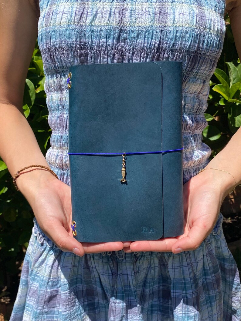 May include: A teal leather journal with a blue elastic closure and a gold charm. The journal has gold corner protectors and the initials "H.A." embossed on the lower right corner. The journal is held in front of a blue and white patterned dress.
