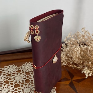 May include: A burgundy leather journal with gold-colored eyelets and charms. The journal is closed with a red cord and features a gold owl charm, a heart charm, and a shell charm. The journal is standing upright on a wooden surface.