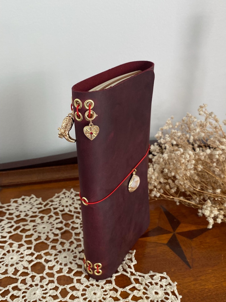 May include: A burgundy leather journal with gold-colored eyelets and charms. The journal is closed with a red cord and features a gold owl charm, a heart charm, and a shell charm. The journal is standing upright on a wooden surface.