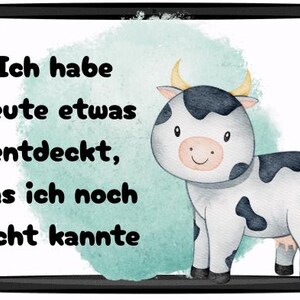 May include: A framed print featuring a watercolor illustration of a cartoon cow with black spots and yellow horns. The text reads "Ich habe heute etwas entdeckt, das ich noch nicht kannte."