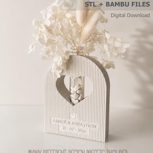 Könnte beinhalten: Eine gerippte, bogenförmige Vase mit einem herzförmigen Ausschnitt in der Mitte, die ein klares Glasröhrchen mit kleinen weißen Herzen enthält. Die Basis zeigt den Text "AIMEE & JONATHON" und "31-07-2026". Text: "heart vase STL + BAMBU FILES".