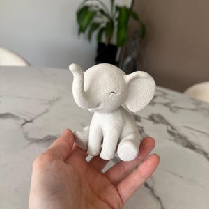May include: A small, white elephant figurine with a textured surface. The elephant is sitting with its trunk raised and eyes closed. The figurine is held in a hand.