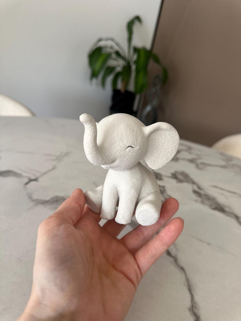 May include: A small, white elephant figurine with a textured surface. The elephant is sitting with its trunk raised and eyes closed. The figurine is held in a hand.
