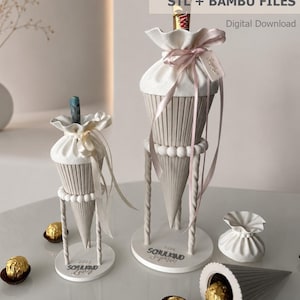 May include: Two 3D printed cone-shaped gift containers with ribbed textures, white fabric-like tops tied with ribbons, and circular bases labeled 'SCHULKIND 2025'. Includes a small separate pouch, a detached cone, and gold-wrapped chocolate candies.