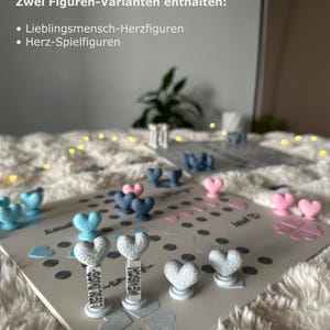 May include: A board game with heart-shaped game pieces in blue, pink, and gray. The game board is light gray with scattered heart and dot decorations. Text on the board reads "Zwei Figuren-Varianten enthalten: Lieblingsmensch-Herzfiguren, Herz-Spielfiguren."
