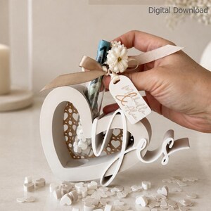 May include: A white 3D-printed heart-shaped container with the script word yes attached to the front. It includes a clear glass tube holding a rolled banknote, decorated with a beige ribbon, a small white flower, and a white tag with handwritten text.