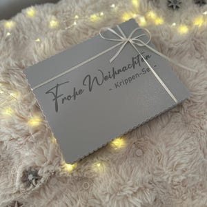 Puede incluir: Una caja rectangular gris con un lazo blanco. La caja tiene la inscripci&oacute;n "Frohe Weihnachten" en una fuente cursiva. Descansa sobre una superficie suave y clara, iluminada por peque&ntilde;as luces. Un regalo.
