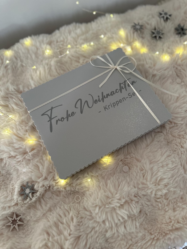 Puede incluir: Una caja rectangular gris con un lazo blanco. La caja tiene la inscripci&oacute;n "Frohe Weihnachten" en una fuente cursiva. Descansa sobre una superficie suave y clara, iluminada por peque&ntilde;as luces. Un regalo.