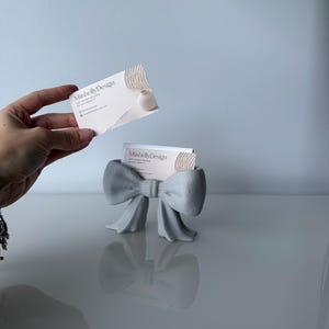 Puede incluir: Un tarjetero gris claro con forma de lazo y superficie texturizada. El soporte contiene una tarjeta de visita blanca con el texto MiishellyDesign, soft, modern 3D prints for your home e iconos de redes sociales. Una tarjeta similar se sostiene arriba.
