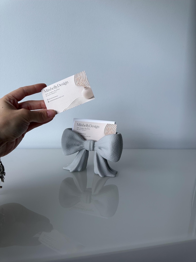 Puede incluir: Un tarjetero gris claro con forma de lazo y superficie texturizada. El soporte contiene una tarjeta de visita blanca con el texto MiishellyDesign, soft, modern 3D prints for your home e iconos de redes sociales. Una tarjeta similar se sostiene arriba.