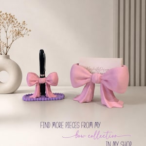 Puede incluir: Un portal&aacute;pices rosa con forma de lazo sobre una base ovalada morada con cuentas y un tarjetero rosa con forma de lazo. El texto dice: FIND MORE PIECES FROM MY bow collection IN MY SHOP. Un bol&iacute;grafo negro est&aacute; insertado en el portal&aacute;pices.