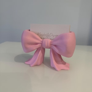 Puede incluir: Un tarjetero en forma de lazo rosa claro con superficie texturizada. El soporte presenta un nudo central y dos extremos colgantes. Una tarjeta de visita rectangular blanca con el texto impreso MushellyDesign 3D Design Studio est&aacute; insertada.