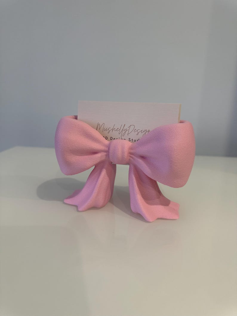 Puede incluir: Un tarjetero en forma de lazo rosa claro con superficie texturizada. El soporte presenta un nudo central y dos extremos colgantes. Una tarjeta de visita rectangular blanca con el texto impreso MushellyDesign 3D Design Studio est&aacute; insertada.