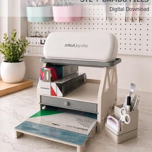 May include: A white Cricut Joy Xtra machine sits atop a beige multi-tiered desk organiser. The unit features a storage shelf, a small drawer, and a bottom tray holding a cutting mat. A separate beige pen and tool holder sits to the right.