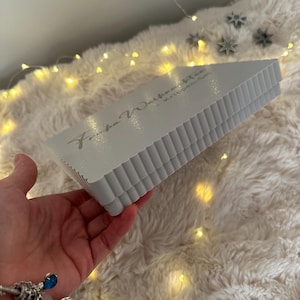 Puede incluir: Una caja triangular gris claro con un borde corrugado se sostiene en una mano. La caja tiene texto. El fondo es una superficie blanca y esponjosa con peque&ntilde;as luces amarillas y decoraciones de estrellas plateadas.