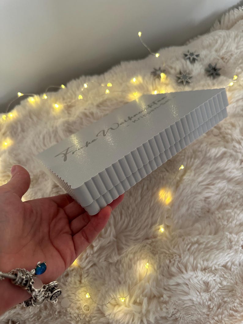 Puede incluir: Una caja triangular gris claro con un borde corrugado se sostiene en una mano. La caja tiene texto. El fondo es una superficie blanca y esponjosa con peque&ntilde;as luces amarillas y decoraciones de estrellas plateadas.