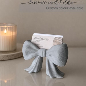 May include: A light grey business card holder shaped like a bow, holding a white business card with the text MiishellyDesign. The text business card holder and Custom colour available appear at the top of the image above the bow object.