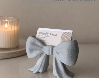 May include: A light gray business card holder shaped like a bow, holding a white business card with the text MiishellyDesign. The text business card holder and Custom colour available appear at the top of the image above the bow object.