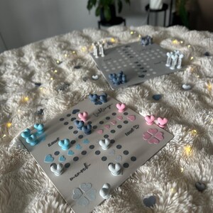 May include: Two gray game boards with heart-shaped and clover-shaped game pieces in blue, pink, and white. The boards are on a fluffy white surface, with a few scattered heart-shaped pieces. The word "Liebling" is visible.