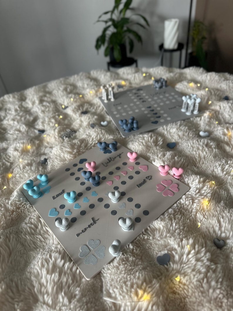 May include: Two gray game boards with heart-shaped and clover-shaped game pieces in blue, pink, and white. The boards are on a fluffy white surface, with a few scattered heart-shaped pieces. The word "Liebling" is visible.
