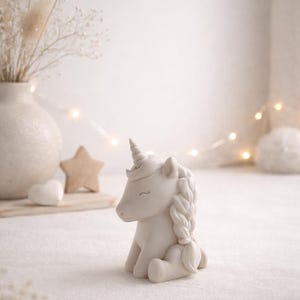 Może przedstawiać: Biała ceramiczna figurka jednorożca z zaplecioną grzywą i spiralnym rogiem. Jednorożec siedzi, z głową obr&oacute;coną na bok. Tło obejmuje wazon z suszonymi kwiatami, gwiazdę i serce.