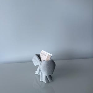 Puede incluir: Un tarjetero de color gris claro con forma de lazo, hecho de un material mate y texturizado. El soporte cuenta con una ranura central que contiene una tarjeta de visita blanca con el texto Mishell Design impreso. El dise&ntilde;o imita un lazo.